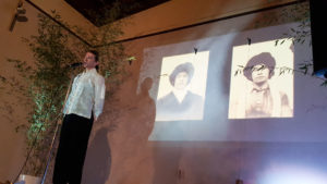 A man stands on a stage in front of a projection of two historical photographs