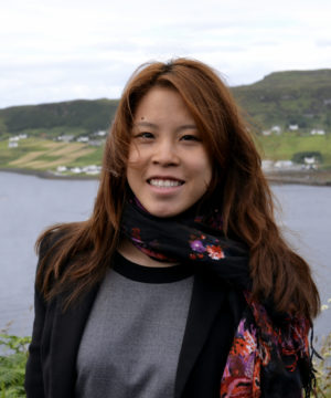 Clarissa Jeakings stands in front of a lake, smiling at the camera. She is wearing a grey shirt, a black blazer and a floral silk scarf.